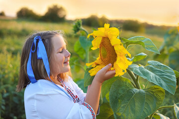 Beautiful girl in embroidered shirt with yellow and blue ribbons in hair with sunflower on field....
