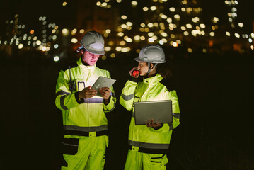 Team of two industrial workers using laptop, walkie-talkie, and phone at night. Represents remote communication, technology integration, and collaborative work in the energy sector