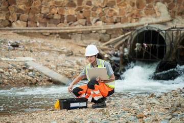 Female engineer with laptop inspecting water drainage pipes at an industrial site. Concept of environmental monitoring, infrastructure management, and technology in engineering