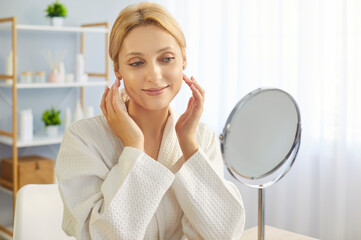 Smiling young woman in bathrobe gently touching her cheeks while sitting at table and admiring her...