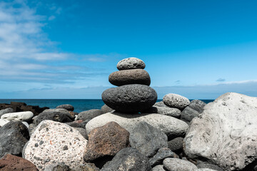 Balanced stack of smooth volcanic stones on a rocky beach, with the deep blue ocean and a partly cloudy sky in the background, evoking peace and stability.