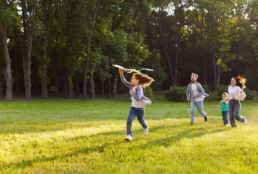 Happy family flying kite in summer park, parents and kids playing together outdoors. Mother, father and children running on green grass of lawn, child holding kite ready to fly. Freedom concept - Powered by Adobe
