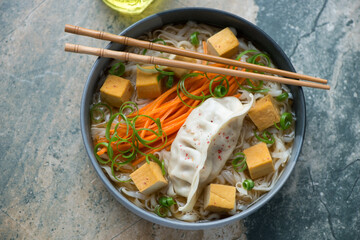 Bowl of rice noodles with smoked tofu and steamed dumpling, horizontal shot on a beige and grey...