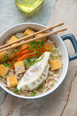 Rice noodles in miso broth with steamed dumpling and tofu, vertical shot on a grey and roseate granite background, elevated view
