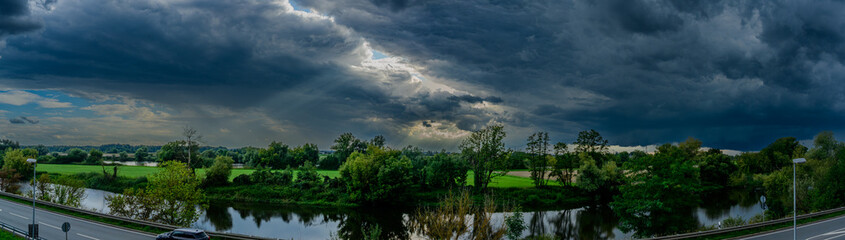 Dramatische Wolkenformation über einem Fluss in der Abenddämmerung mit grünen Ufern und...