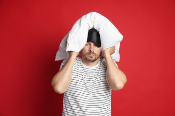 Man with pillow and sleep mask suffering from hangover on red background