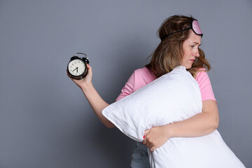 Woman with pillow and alarm clock suffering from hangover on grey background