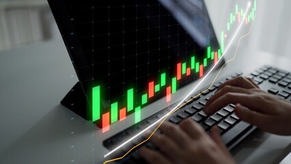Hands typing on a keyboard in front of a screen displaying a stock market graph. The image captures...