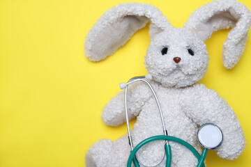 Pediatrics concept. Toy bunny with stethoscope on yellow background, top view. Space for text
