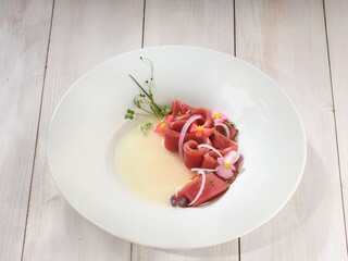 Thin slices of red tuna are arranged gracefully in a white bowl with a shallow pool of pale yellow sauce, adorned with tiny flowers, herbs, and slivers of white onion