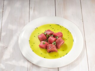 Several small rolls of rare tuna sit nestled in a bright green sauce, garnished with fresh greens, presented on a white plate. A bright atmosphere and white table complete the dish
