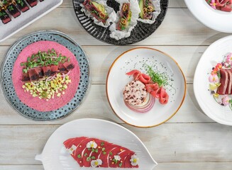 Six plates of diverse seafood appetizers are beautifully arranged on a white wooden table. Each...