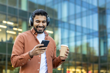 A smiling man enjoys music and coffee while using his phone in front of a modern building.