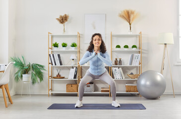 Yogi sporty fit woman performing squat on yoga mat at home studio gym room, exercising with energy, fitness and wellness, reflecting positive emotions in domestic situation, personal wellness practice