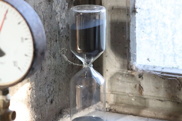Close-up of a black-sand hourglass in a dusty, abandoned industrial building. Natural light from the window highlights the contrast between time, decay, and forgotten machinery.