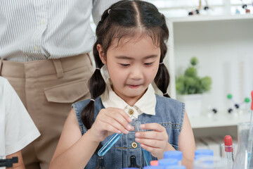Asian elementary girl using pipette carefully mixing blue liquid in beaker while participating in classroom science experiment with microscope and lab tools placed on table during chemistry lesson