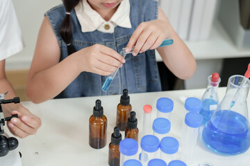 Closeup of asian girl using pipette to pour blue liquid into test tube while conducting science experiment on white lab desk filled with glass bottles and classmates around