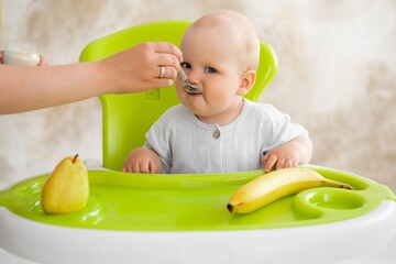 feeding a baby, mother feeding a baby boy with a spoonful of fruit puree in a green high chair, baby food