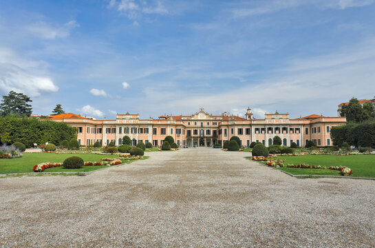 Varese, Italy: The grand Palazzo Estense, a historic palace with a pale facade, set amidst a meticulously manicured Baroque garden under a blue, cloudy sky.