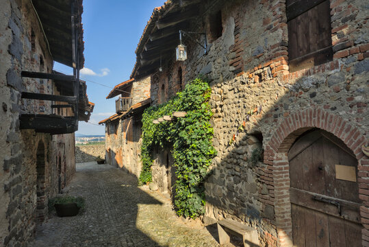 Ricetto di Candelo, Italia: Una stretta strada di ciottoli nel borgo medievale con muri in mattoni e pietra coperti di edera, illuminata dal sole sotto un cielo azzurro.