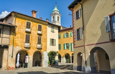 Biella, Italy: A sunlit, historic cobblestone square surrounded by colorful buildings with archways...