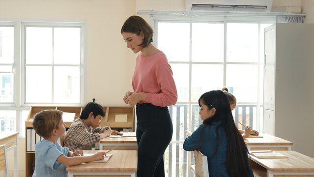 Caucasian teacher walking and check student homework at classroom while diverse children doing classwork at classroom. Diverse smart student lecturing and taking a note at elementary school. Pedagogy.