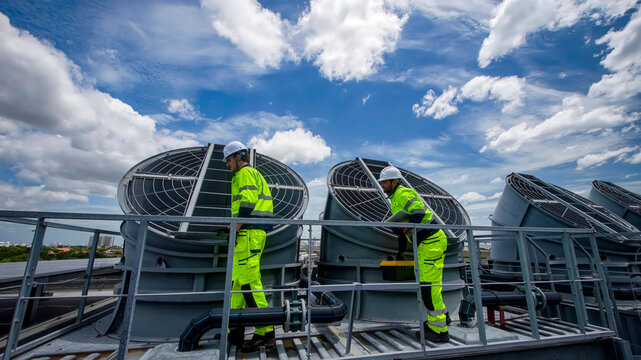 Workers perform maintenance on rooftop ventilation units under a cloudy sky
