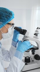 Female scientist wearing blue protective gear using a microscope examining samples in a laboratory...