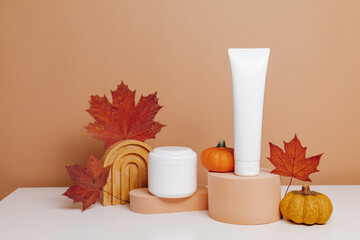 Mockup facial cream with bottle of body lotion on podium with autumn leaves and pumpkins on brown background, seasonal skincare