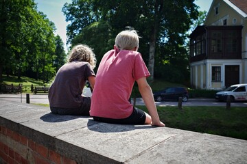 Two boys sitting on a brick wall on a sunny day, relaxing and enjoying the outdoors near a residential area with trees and parked cars in the background.