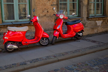 Two bright red scooters parked on a cobblestone street in front of an old weathered building with green-framed windows, urban transportation and retro style concept.