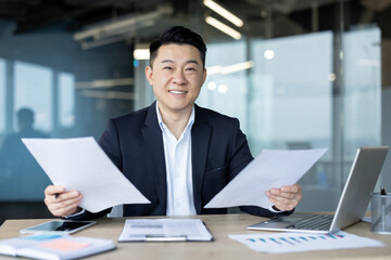 Portrait of a young Asian businessman working in the office with a laptop and documents, sitting at a desk, holding papers and looking at the camera