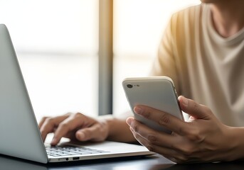 Medium Shot of Adult Using Silver Laptop and Holding White Smartphone on Dark Reflective Table in Bright Indoor Office Setting