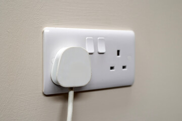 White UK 3-pin mains plug inserted into modern dual wall socket with switches. Mounted on a light...