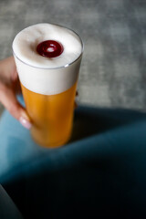 Close-up of a hand holding a tall glass of beer with thick foam and a red candy on top, against a...