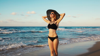 Fototapeta premium Summer vacation, beautiful happy slim woman in black swimsuit, tourist hat on the beach on sea