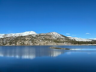 June Lake, California. Expansive mountain lake with perfect reflections under brilliant blue sky. Snow-dusted peaks frame the water while evergreen trees dot the surrounding hills and shoreline