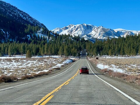 Straight highway stretching toward snow-covered mountains under clear blue sky, June Lake, California. Red SUV drives down center of empty road through winter landscape with scattered snow