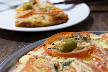 Part of an Argentinian Neapolitan pizza on a tray and a slice on a plate in the out-of-focus background