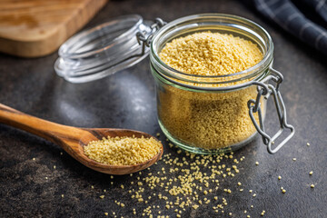 Raw couscous in spoon on black table.