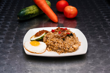 Kampong Nasi Goreng with Fragrant Fried Rice, Fried Egg, Sambal, Cucumber and Garnishes on a White Plate isolated on dark background side view of Malaysian food