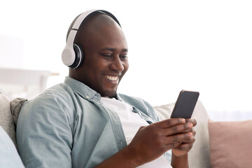 Relaxed middle aged african american man listening music on smartphone with wireless headphones, enjoying favorite songs on mobile phone, resting on comfy sofa in living room, free space