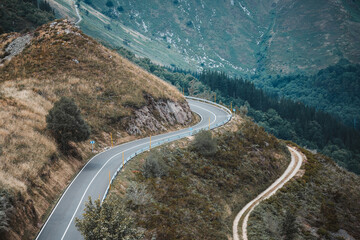 Telephoto view of a winding asphalt mountain road with safety railings, flanked by a rugged hiking...