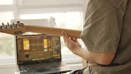 Musician composing music at home, working on laptop with DAW and playing the blue electric guitar. Creative music production process in cozy daylight home studio.