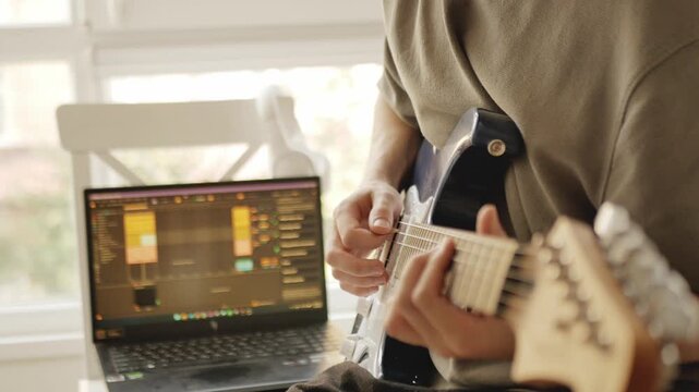 Guitarist performing electric guitar solo while recording music in cozy home studio with laptop and professional audio software. Music performance, creativity, songwriting