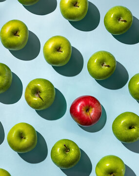 One red apple standing out in a group of green apples on a blue background. Concept of diversity, individuality, inclusion, uniqueness, and thinking differently. Minimalist top view.