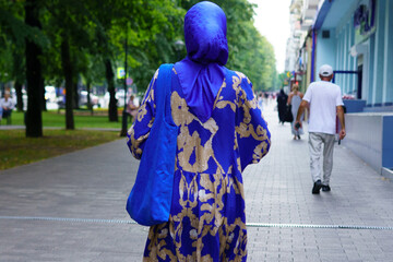 Colorful hijab-clad woman walks along a busy urban street surrounded by trees on a sunny day