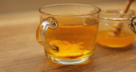 Hot tea with slice of lemon rotating in transparent cup on wooden table. Honey in glass jar with honey stick complements warm beverage with natural sweetness