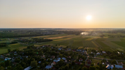 Aerial view of a serene countryside landscape during sunset, showcasing fields, scattered houses, and a warm golden hue reflecting off the horizon.