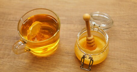 Transparent cup of aromatic tea with fresh lemon slice and honey jar stand on wooden table. Fragrant beverage brews in glass mug enriched with sour fruit